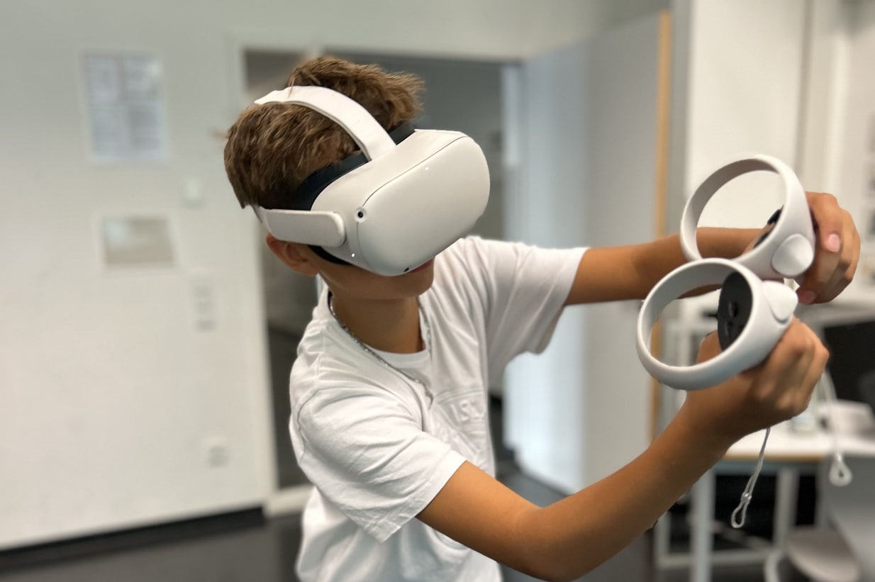 Ein Junge trägt eine VR-Brille und hält zwei VR-Controller in einer modernen Lernumgebung. Er ist in ein interaktives Virtual Reality Erlebnis vertieft, das vermutlich Teil eines Bildungsprogramms ist. Der Junge trägt ein weißes T-Shirt und befindet sich in einem hellen Raum mit klaren Wänden und Möbeln im Hintergrund. Das Bild zeigt die Nutzung innovativer Technologien zur Förderung von Lernen und Engagement bei Jugendlichen.