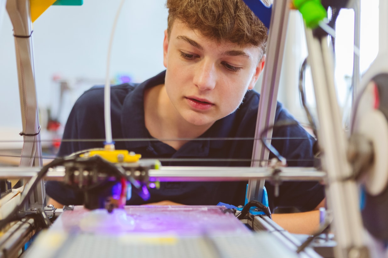 Ein junger Mann in einem dunkelblauen Poloshirt beobachtet aufmerksam den Arbeitsprozess eines 3D-Druckers. Der 3D-Drucker ist aktiv und druckt ein Objekt, während der Junge sich über das Gerät beugt, um den Fortschritt genau zu verfolgen. Der Hintergrund deutet auf eine technische Werkstatt oder ein Labor hin, was auf eine Lernumgebung im Bereich moderner Fertigungstechniken hinweist. Das Bild vermittelt den Einsatz fortschrittlicher Technologien zur Förderung technischer Fähigkeiten und praktischen Wissens bei Jugendlichen.